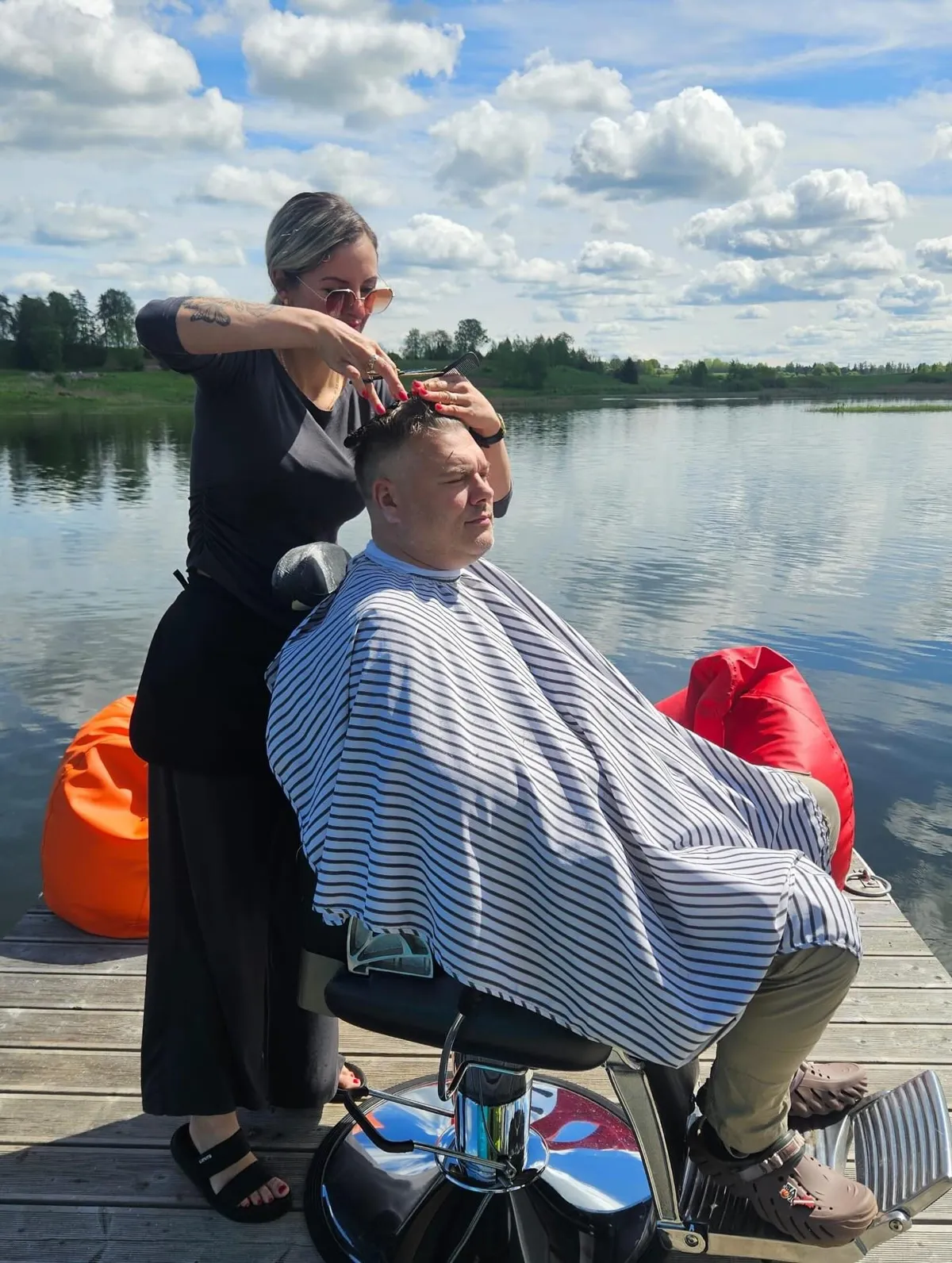 Frizieris griež klienta matus uz plosta pie ezera saulainā dienā